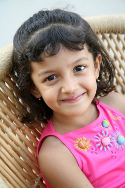 6 yıllık küçük bir kız hasır sandalyede oturuyor ve gülümsüyor.