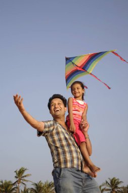 Güney Asyalı Hintli kız babasının omzunda oturmuş deniz kıyısında renkli uçurtmalar uçuruyor. 