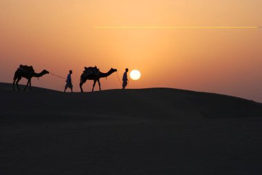 Setting sun ahead of two camel men in desert, Khuri, Jaisalmer, Rajasthan, India  clipart