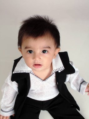 Güney Asyalı 7 aylık bebek tişört, ceket ve pantolon giyiyor oturuyor ve kameraya doğru bakıyor.