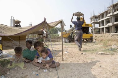 İnşaat işçilerinin çocukları geçici bir barınak altında oynarken ebeveynleri sıcak öğleden sonra güneşi, Ahmedabad, Gujarat, Hindistan 'da çalışıyor. 