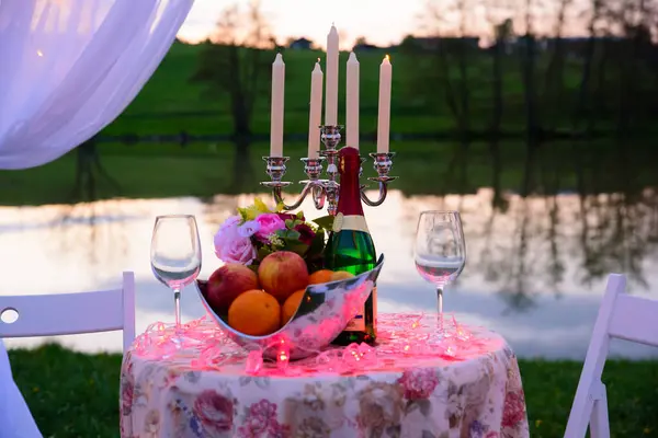 A romantic place for a date or marriage proposal in a park on the lake, in a beautiful tent and decorated with candles and garlands. A table set with fruit and a bottle of wine.
