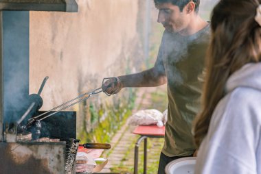 Siyah saçlı ve yeşil tişörtlü genç bir adam barbeküde maşayla et pişiriyor..