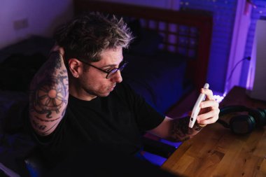 Genç adam geceleri odasında sandalyesinde otururken telefonuyla video izliyor..