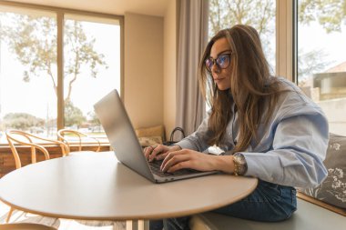 Açık mavi gömlekli ve gözlüklü girişimci kadın laptopuyla konsantre bir şekilde çalışıyor..