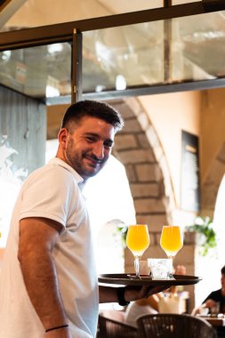 Beyaz polo tişörtlü garson. Bir elinde iki bira tepsisi, bir elinde gülümseme. Dikey.