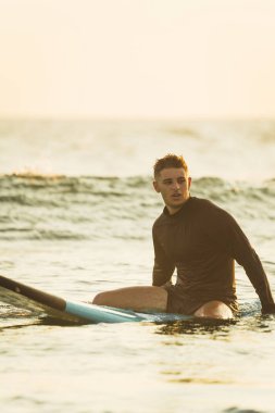 Gün doğumunda sörf tahtasında oturan bir sörfçü..