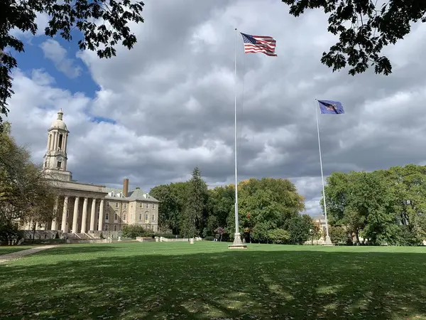 State College, Pennsylvania, ABD - 7 Ekim 2023: State College 'deki Pennsylvania State University kampüsündeki eski ana bina, Amerika Birleşik Devletleri bayrağı ve önünde yapraklarla çerçevelenmiş Pensilvanya bayrağı.