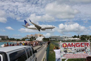 Sint Maarten, Netherlands, November 26, 2016: Airplane lands at Princess Juliana International Airport, in front of busy street and crowded beach. The airport has very low-altitude flyover landing approaches because the runway is close to the shore. clipart