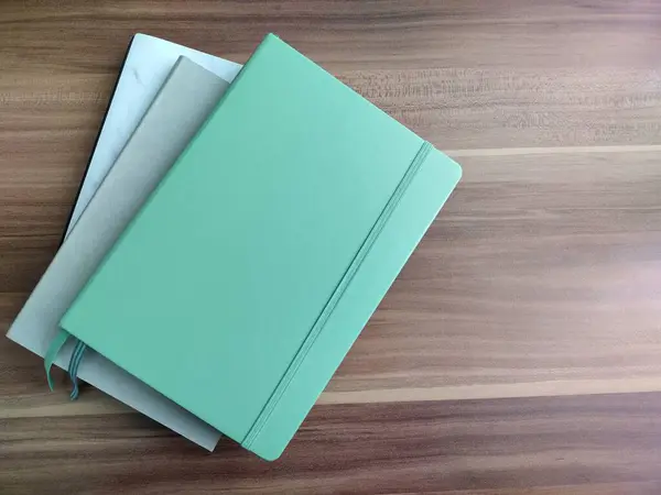 Beyaz mermer bir defter, lastik bantlı yeşil bir not defteri ve tahta bir masaya yaslanmış gri bir günlük. Kuş görünümü.