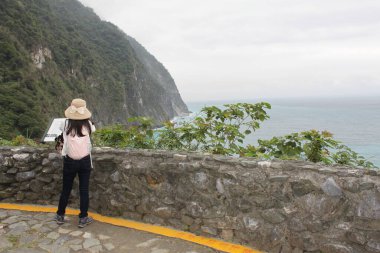 Hualien, Tayvan-Mart 18, 2024: Uzun siyah saçlı Asyalı kadın Quingshui uçurumundaki kaya korkuluklarında sırt çantası ve güneş şapkası takan sahile bakıyor. Hualien, Tayvan 'da Pasifik Okyanusu. Tropik kıyılar, ada. 
