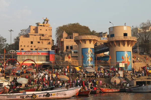 stock image Varanasi, India- March 13, 2019: Scene of daily life at Dr. Rajendra Prasad Ghat, at the banks of river Ganges in Varanasi at sunrise. Crowded with boats and people. Big wall paintings of Hindu Gods Parvati and Lord Shiva at two towers.