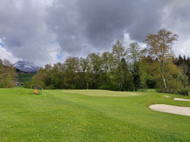 Bad Aussee 'de iyi bakılmış golf sahası, ilkbaharda Alpler' de Avusturya: yeşil çayırlar, çiçek açan çiçekler, ağaçlarda yeşil yapraklar ve arka planda karlı dağ zirvesi. Renkli bayrak direği, kum sığınakları, çimenler.