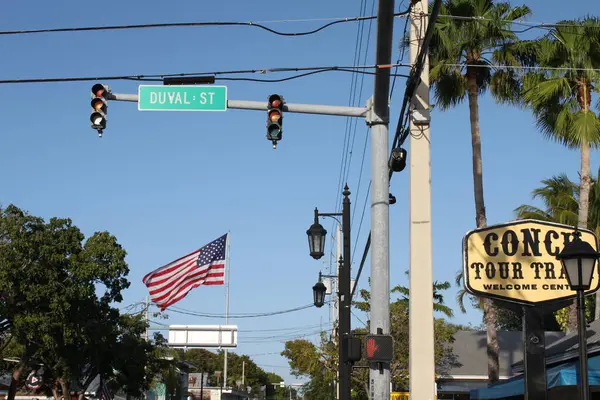 Key West, Florida, ABD - 12 Şubat 2020: kavşakta Duval caddesi yazan tabela, büyük Amerikan bayrağı sallama, fenerler ve trafik ışıkları. Deniz kabuğu turu tren karşılama merkezinin büyük işareti.. 