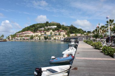 Marigot, Saint Martin, France- November 26, 2016: View of wooden pier, dinghy with motor, residences at shore of carribean sea, palmtrees, flags and castle. Waterfront at Marigot. clipart