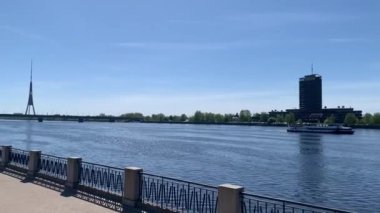Riga, Letonya, 3 Mayıs 2024: River Daugava, Riga Radyo ve TV Kulesi 'ndeki işgal edilmiş tekne görüntüsü, gezinti güvertesinde yürüyen insanlar, Letonya bayrağı sokakta dalgalanıyor. Kamera demiryolu köprüsüne geri döndü.