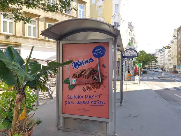 stock image Vienna, Austria, Juli 5, 2024: Advertisement for traditional, famous austrian sweets, cream filled wafers, produced by company Manner. Bus stop in Vienna. Sunny day, framed by plants, historic houses.