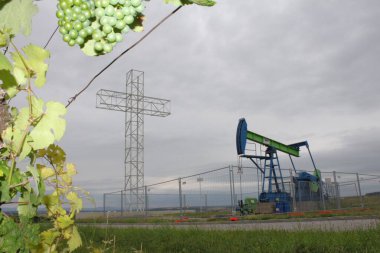 Matzen, Gnserndorf, Lower Austria - July 25, 2010: OMV oil pumpjack, vine and miners monument Barbara cross. Austrian region Weinviertel. Framed by grapevine with leaves and green grapes. Grey Sky clipart