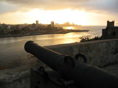 Old Havana, Havana, Cuba - March 15, 2009: Fortress Castillo del Morro guarding the entrance to Havana harbor. Cannon on wooden carriage aiming over merlons from massive walls. Evening view to city center with Malecon, sunset. clipart