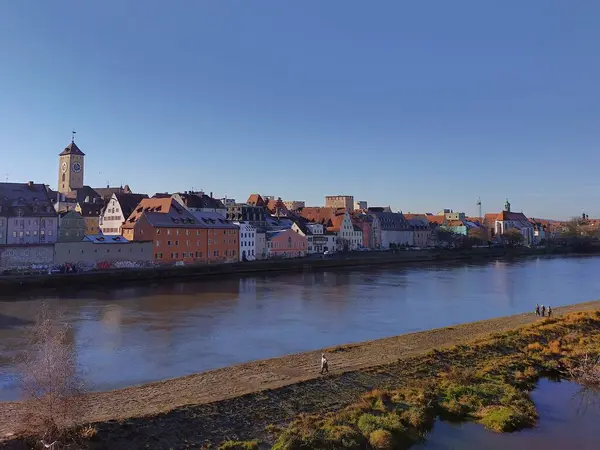 Regensburg, Bavyera, Almanya, 1 Aralık 2024: Liman manzarası ve eski Regensburg kasabası. Tarihi, renkli binalar, kule, Tuna nehri. Nehir kıyısında yürüyen insanlar. Kışın güneşli bir gün.