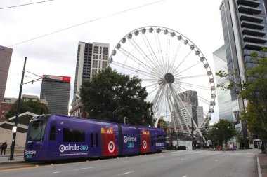 Atlanta, GA, USA - September 28, 2024: View of giant Ferris Wheel in downtown Atlanta, Georgia in Centennial Olympic Park. Purple public tram (light rail vehicle, popular streetcar) stops in front. clipart