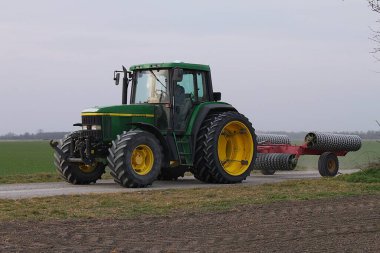 Kopfstetten, Gaenserndorf, Lower Austria - March 08, 2025: John Deere 6610 tractor with dual wheels on rear axle pulls ridged roller on dirt road. clipart