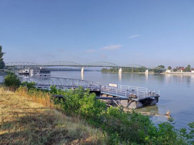 Esztergom, Macaristan, 5 Ağustos 2024: Gezinti güvertesindeki Tuna Nehri rıhtımının manzarası. Nehir turları için popüler bir durak. Güneşli bir yaz sabahı. Arka planda Slovakya 'ya giden sınır köprüsü.