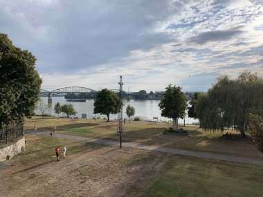 Esztergom, Macaristan, 4 Ağustos 2024: Tuna nehrinin kıyısında güneşli bir öğleden sonra. İnsanlar eğleniyor, yürüyüşe çıkıyorlar. Nehir yolcu gemisi köprünün altından geçiyor..