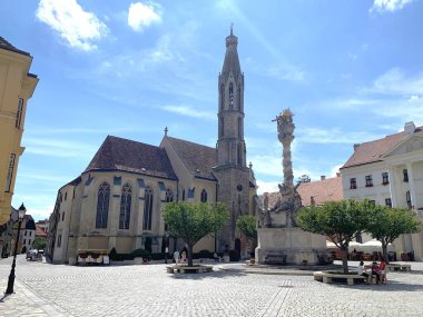 Sopron, Macaristan, 9 Ağustos 2024: Kutsanmış Mary Benedictine Kilisesi (Macarca: Kecske templom) ve Sopron 'daki Kutsal Üçleme Heykeli (Foter). Güneşli yaz günü.