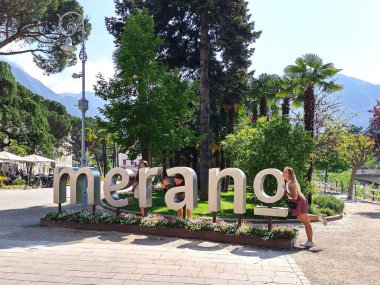 Meran, İtalya 2 Mayıs 2025: Güney Tyrol 'de Merano veya Meran kentinin büyük harfli dekoratif cazibesi. Kuzey İtalya. Genç turistler ikonik tabelayla fotoğraf çekiyorlar. Çiçek süslemesi. Seyahat teması