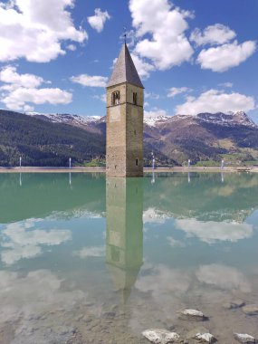 Alt Graun (Vecchio Curon), Vinschgau, Trentino, South Tyrol, İtalya - 1 Mayıs 2025: Reschen Gölü 'ndeki (Reschensee, Lago di Resia) su altındaki kilise kulesinin dikey görünümü. Tam bir yansıma. Güzel bir dağ manzarası. Baharda güneşli bir gün.