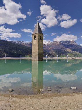 Alt Graun (Vecchio Curon), Vinschgau, Trentino, South Tyrol, İtalya - 1 Mayıs 2025: Reschen Gölü 'ndeki (Reschensee, Lago di Resia) su altındaki kilise kulesinin dikey görünümü. Sudaki yansıma. Güzel bir dağ manzarası. Baharda güneşli bir gün.