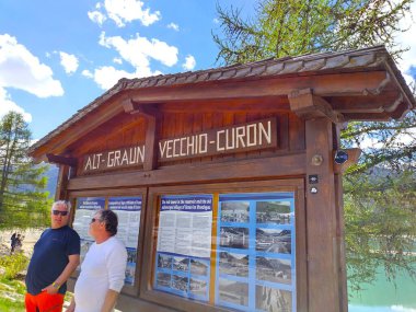 Alt Graun (Vecchio Curon) Vinschgau, İtalya - 1 Mayıs 2025: Suyun altındaki Graun köyü hakkında bilgi verir. Gölgede duran iki erkek turist. Baharda güneşli bir gün.
