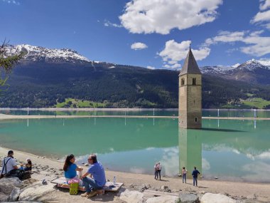 Alt Graun (Vecchio Curon), Vinschgau, Trentino, Güney Tyrol, İtalya - 1 Mayıs 2025: Reschensee gölündeki turistler (Lago di Resia, Lake Reschen) batık 14. yüzyıl kilise kulesini keşfediyorlar. Baharda güneşli bir gün.