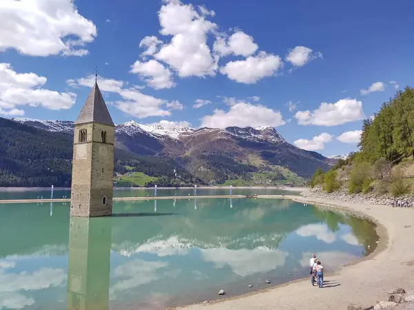 Alt Graun (Vecchio Curon), Vinschgau, Trentino, South Tyrol, İtalya - 1 Mayıs 2025: Reschensee (Lago di Resia Gölü, Reschen) nehir kıyısında yürüyen turistler. Batık kilisenin çan kulesini ziyaret ediyorum. Baharda güneşli bir gün