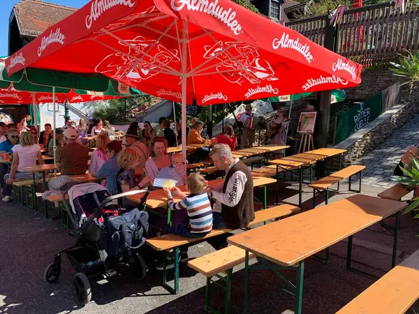 Viyana, Avusturya - 24 Ağustos 2025: Bir aile, geleneksel Viyana şarap üreticisi festivali Neustifter Kirtag sırasında bir şarap tavernasının gölgeli açık hava yemek alanında oturuyor. Arka planda canlı müzik.