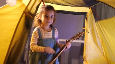 Küçük kız evdeki bir çadırda otururken gitarla müzik çalıyor. Mutlu bir çocuk şarkı söyler ve eğlenir. Müzikal bir çocuğun ev eğlencesi ya da hobisi. Yüksek kalite 4k görüntü