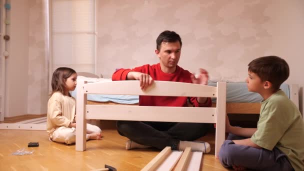父亲和孩子们正在家里组装新家具 孩子们帮爸爸组装床 男孩和女孩帮助父母做家务活 — 图库视频影像