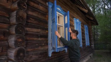 Eski ahşap bir evin penceresinin panjurlarını açan bir adam. Belarus kırsalındaki bir evde ahşap panjurların açılması. Doğu Avrupa 'daki kır evi