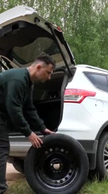 Yol kenarındaki bir arabanın patlak lastiğini değiştiren dikey bir video. Şoförün kendisi patlak lastiği yedek lastikle değiştiriyor. Şoför arabanın bagajındaki yedek lastiği çıkardı.