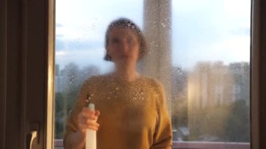 Genç bir kadın pencere camına deterjan sıkar. Bir kadın pencereyi temizler. Bir kadın ev işlerini yapar ve evi temizler..
