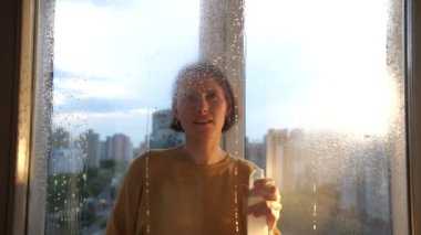 Genç bir kadın bir pencere camına deterjan püskürtür ve bir bez parçasıyla kurutur. Bir kadın dairesinde bir pencereyi yıkıyor. Kadın gülümsüyor ve ev işlerini zevkle yapıyor, daireyi temizliyor.