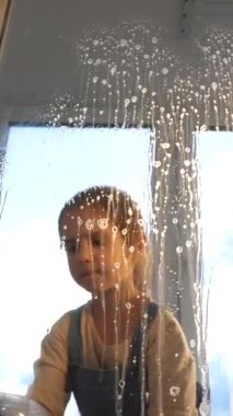 Dikey videocu kız pencere camını bir bezle siliyor. Küçük kız pencereyi temizliyor. Küçük bir kız daireyi temizliyor ve ev işlerini yapıyor. alt görünüm