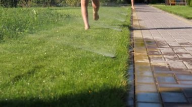 Otomatik fıskiyeyi çalıştırırken çimlerde koşan çocuklar. Çocuklar yazın suyla oyun oynarlar. Sıcak bir gün. Çimenlik fıskiyesinin altında serinleyen bir çocuk.