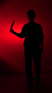 Vertical video mans silhouette standing against a stark red background, holding a sharp knife, creating a chilling atmosphere of danger, crime, and threat