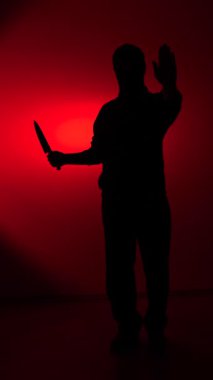 Vertical video dark silhouette of a person holding a large knife with raised hand against harsh red light conveying threat, fear, and violent menace