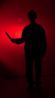Vertical video shadowed figure of a person standing against a dark red background, holding a sharp kitchen knife in a threatening manner, conveying concepts of danger, crime, and halloween horror