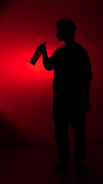 Vertical video mans full body silhouette with a bottle of alcohol, drinking against a dark red background, symbolizing addiction, despair, and the struggles of alcoholism