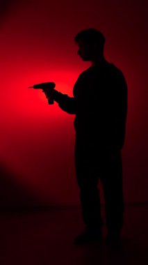 vertical video Man silhouette holding a power drill, creating a menacing and dangerous mood under a bright red spotlight, emphasizing anonymous threat and hidden intent