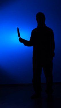 vertical video Person silhouette standing in a menacing posture, holding a large knife, creating a scary and dangerous atmosphere, with a blue light creating a moody background